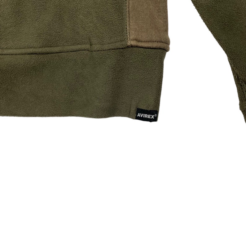 Puede incluir: Primer plano de una sudadera de forro polar verde oliva con un panel marr&oacute;n en la parte superior derecha. El dobladillo inferior presenta una etiqueta negra con el nombre de la marca "AVIREX" en blanco.