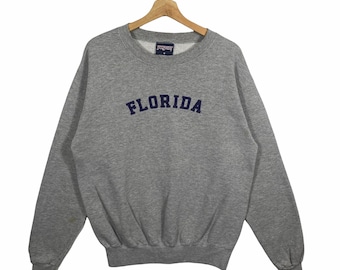 Puede incluir: Sudadera gris con un logotipo "FLORIDA" azul marino en la parte delantera. La sudadera tiene cuello redondo y mangas largas.