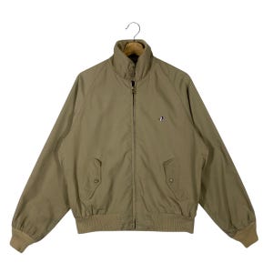 May include: Tan zip-up jacket with a small embroidered logo on the chest. The jacket has a stand-up collar, two front pockets, and ribbed cuffs and hem. The jacket is hanging on a wooden hanger.