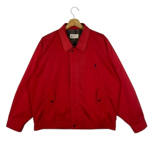 Puede incluir: Chaqueta roja brillante con cierre de botones, dos bolsillos delanteros y cuello. La chaqueta tiene un pequeño logotipo azul marino en el pecho y un forro a cuadros visible en el cuello. Colgada de una percha de madera.