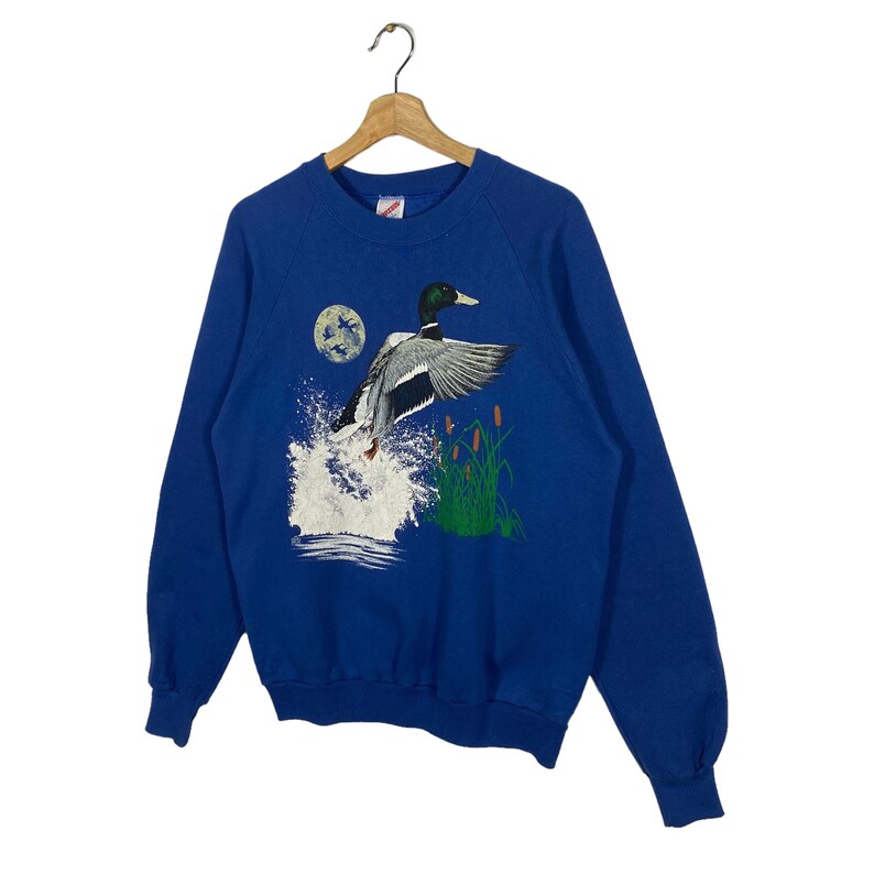 K&ouml;nnte beinhalten: Ein blaues Sweatshirt mit einem Aufdruck eines Enten, das &uuml;ber einen See mit Schilf fliegt. Die Ente befindet sich in der Mitte des Aufdrucks und blickt nach rechts. Der See ist blau und das Schilf ist gr&uuml;n.