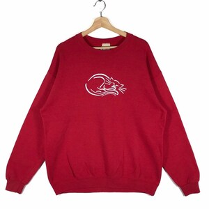 Puede incluir: Sudadera roja con un contorno blanco de un gato durmiendo en la parte delantera. El gato está enroscado y tiene los ojos cerrados. La sudadera tiene cuello redondo y mangas largas.