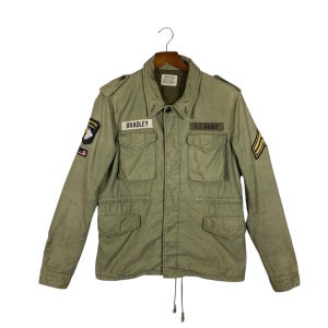 May include: An olive green military jacket with multiple pockets and patches. The jacket features the name "BRADLEY" above the left chest pocket and "U.S. ARMY" above the right chest pocket. The jacket has a button-up front and a drawstring at the waist.