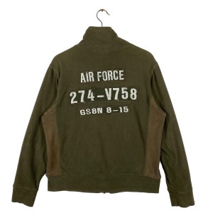Puede incluir: Chaqueta con cremallera verde oliva con paneles laterales marrones. La parte trasera presenta texto blanco que dice "AIR FORCE", "274-V758" y "GS8N 8-15". La chaqueta est&aacute; colgada de una percha met&aacute;lica.