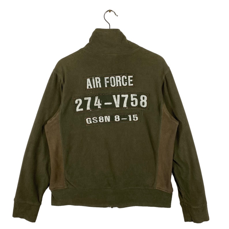 Puede incluir: Chaqueta con cremallera verde oliva con paneles laterales marrones. La parte trasera presenta texto blanco que dice "AIR FORCE", "274-V758" y "GS8N 8-15". La chaqueta est&aacute; colgada de una percha met&aacute;lica.