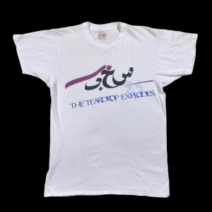 Può includere: Maglietta bianca con una grafica viola e blu di testo arabo che recita "Shujji" e il testo "THE TEARDROP EXPLODES."