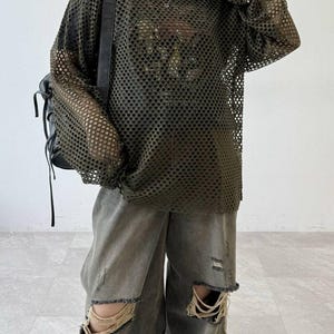 May include: An olive green long-sleeve mesh top with a wide-hole pattern worn over a dark shirt, paired with distressed grey denim jeans featuring large frayed holes at the knees and a black backpack with thin straps worn over one shoulder.