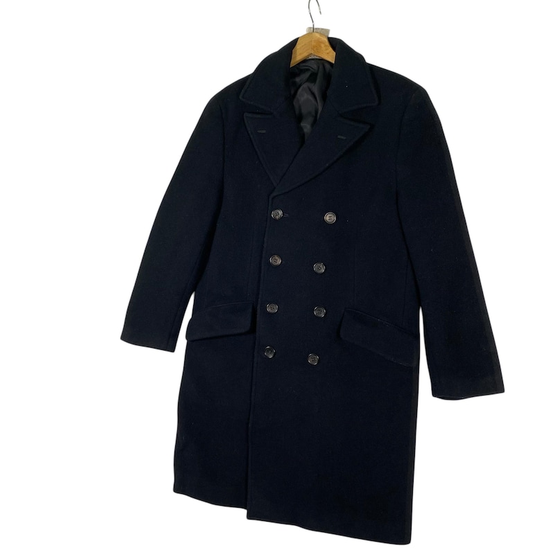 Pu&ograve; includere: Un cappotto doppiopetto blu navy dal design classico. Il cappotto presenta un risvolto a lancia, due tasche anteriori e sei bottoni scuri. Il cappotto &egrave; appeso a una gruccia di legno.