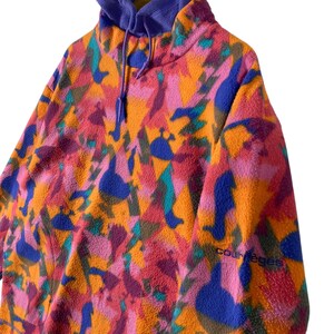 May include: A colorful fleece pullover with a high neck and a drawstring hood. The fleece is a bright orange with pink, blue, and green abstract patterns. The text "Courage" is visible on the left sleeve.