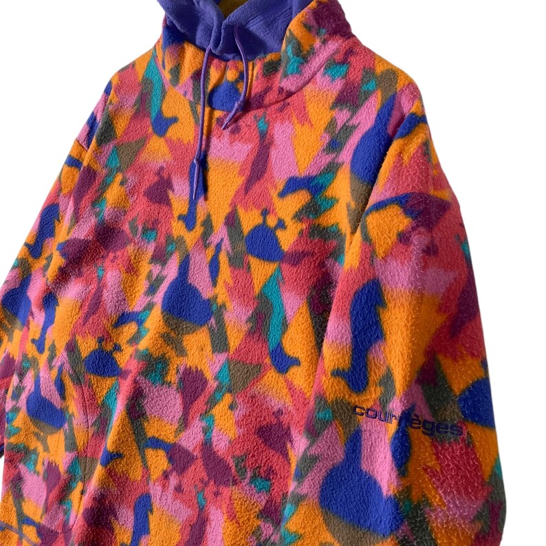 May include: A colorful fleece pullover with a high neck and a drawstring hood. The fleece is a bright orange with pink, blue, and green abstract patterns. The text "Courage" is visible on the left sleeve.