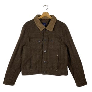 Puede incluir: Chaqueta de lana marrón con cuello de borreguillo beige. La chaqueta tiene dos bolsillos en el pecho, dos bolsillos laterales y cierre de botones. La marca "GAP" es visible en el cuello. Colgada de una percha de madera.