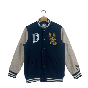 May include: Black varsity jacket with brown sleeves and white stripes. The jacket has a large "D" on the left side and a dog with a collar on the right side. The back of the jacket has a small "3" on the right sleeve.