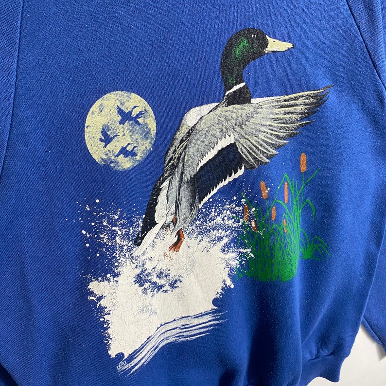 K&ouml;nnte beinhalten: Ein blaues Sweatshirt mit einem wei&szlig;en und grauen Bild eines Enten, das &uuml;ber eine Welle fliegt. Die Ente befindet sich in der Mitte des Bildes und blickt auf die rechte Seite des Bildes. Die Ente fliegt &uuml;ber eine Welle, die hochspritzt. Im Hintergrund des Bildes befindet sich ein Vollmond.