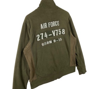 Puede incluir: Chaqueta con cremallera verde oliva con cuello alto. La parte trasera presenta parches con el texto "AIR FORCE", "274-V758" y "GS8N 8-15" en blanco. La chaqueta tiene paneles marrones en los laterales.