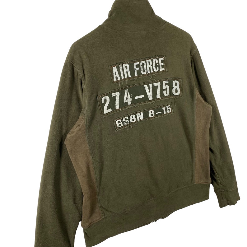 Puede incluir: Chaqueta con cremallera verde oliva con cuello alto. La parte trasera presenta parches con el texto "AIR FORCE", "274-V758" y "GS8N 8-15" en blanco. La chaqueta tiene paneles marrones en los laterales.
