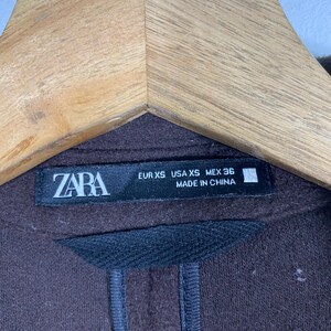 Pu&ograve; includere: Un maglione marrone Zara con un'etichetta nera che recita "ZARA EUR XS USA XS MEX 36 MADE IN CHINA".