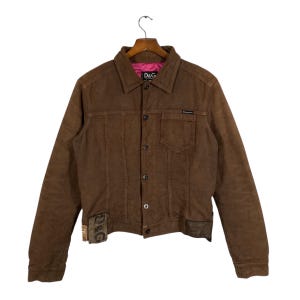 Puede incluir: Chaqueta de pana marrón con botones, cuello y bolsillos. La chaqueta tiene un forro rosa y una pequeña etiqueta D&G. La chaqueta está colgada de una percha de madera.