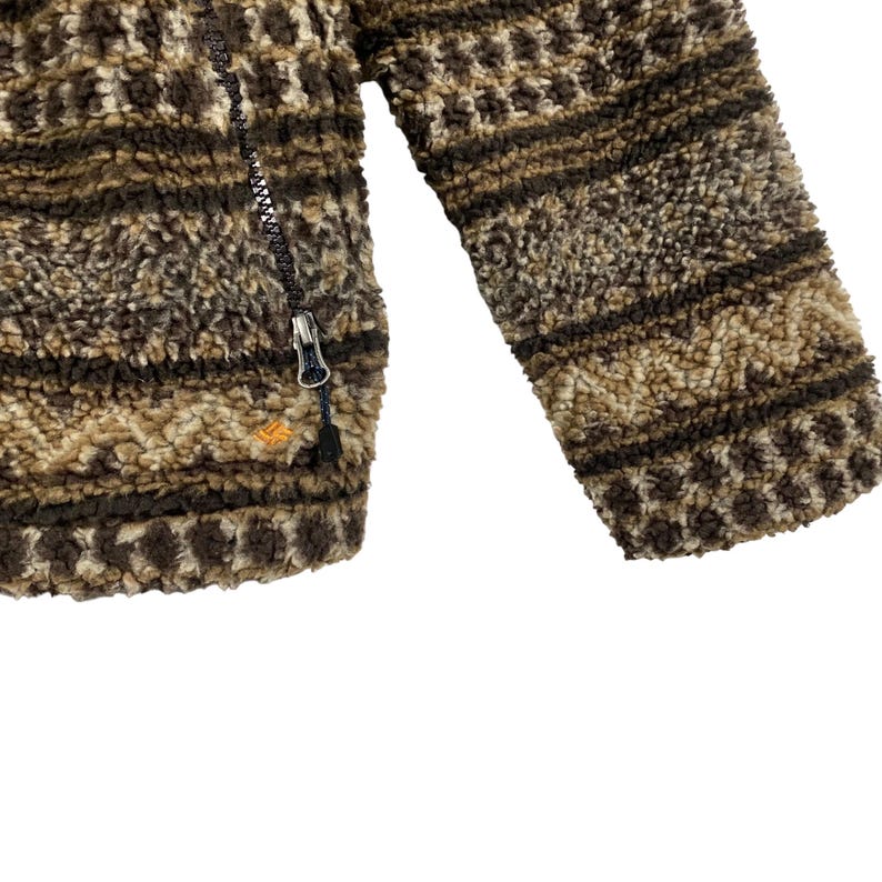 Puede incluir: Una chaqueta de forro polar con estampado marr&oacute;n y beige con cierre de cremallera. La chaqueta tiene una textura peluda y un patr&oacute;n geom&eacute;trico.