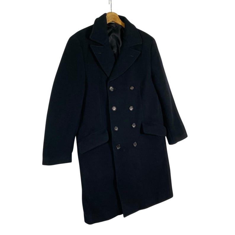 Pu&ograve; includere: Un cappotto nero doppiopetto dal design classico. Il cappotto presenta un colletto a revers, due tasche anteriori e diversi bottoni. Il cappotto &egrave; appeso a una gruccia di legno.