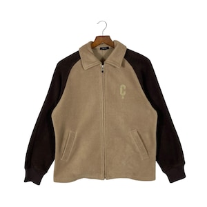 May include: A tan fleece jacket with brown sleeves and collar, hanging on a wooden hanger. The jacket has a full zip, two front pockets, and a small embroidered "C" logo on the chest.