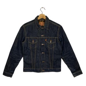 May include: Dark blue denim jacket with button closures, two chest pockets, and a classic collar. The jacket features orange stitching and a small orange tag. It is hanging on a wooden hanger.