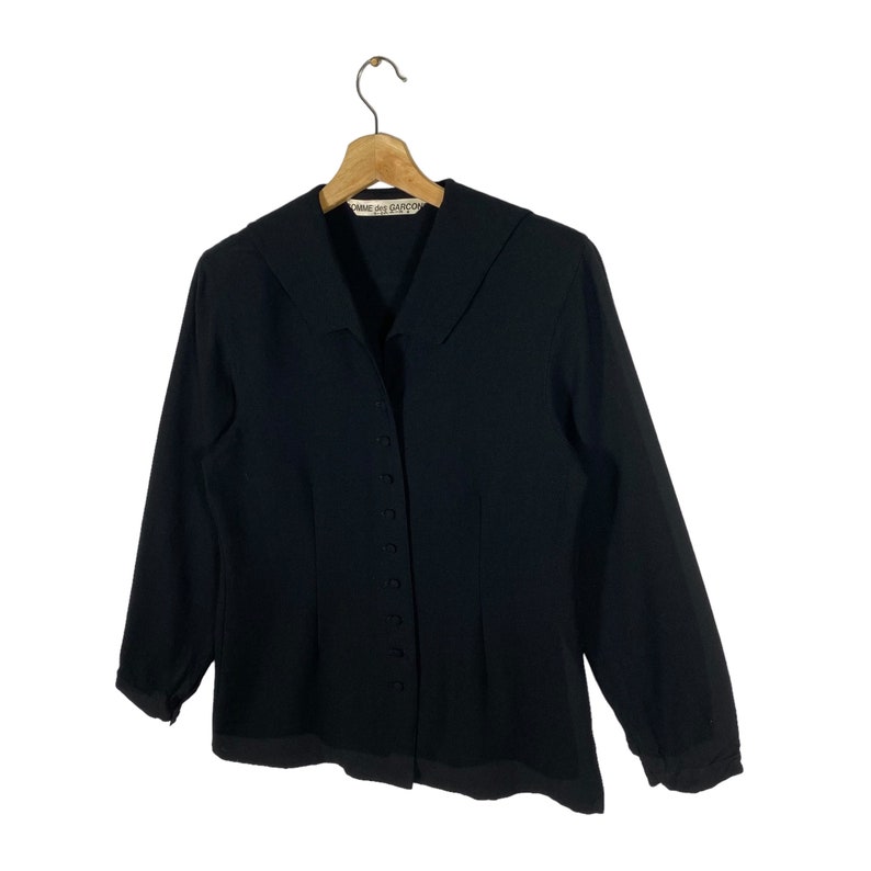 Puede incluir: Una chaqueta negra de botones con cuello y mangas largas. La etiqueta "Comme des Gar&ccedil;ons" es visible en el cuello.