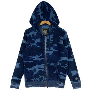 Puede incluir: Una sudadera con capucha azul de camuflaje con un gran logotipo de estrella en la parte delantera. La sudadera tiene una capucha con cordón y un bolsillo canguro.