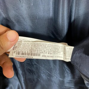 May include: A close-up of a tag with text and numbers. The tag is white with black text and is partially obscured by a dark blue fabric. The text includes company information and product details.