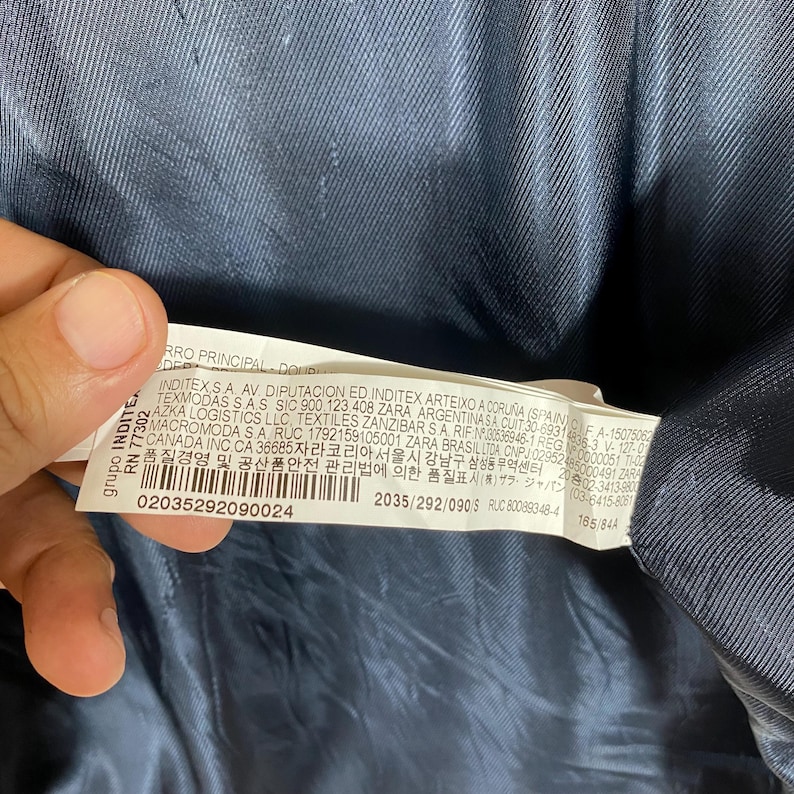 May include: A close-up of a tag with text and numbers. The tag is white with black text and is partially obscured by a dark blue fabric. The text includes company information and product details.