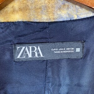 May include: Close-up of a navy blue blazer lining with a ZARA label. The label includes size information and the text "MADE IN MOROCCO". The blazer's interior fabric has a subtle textured pattern.