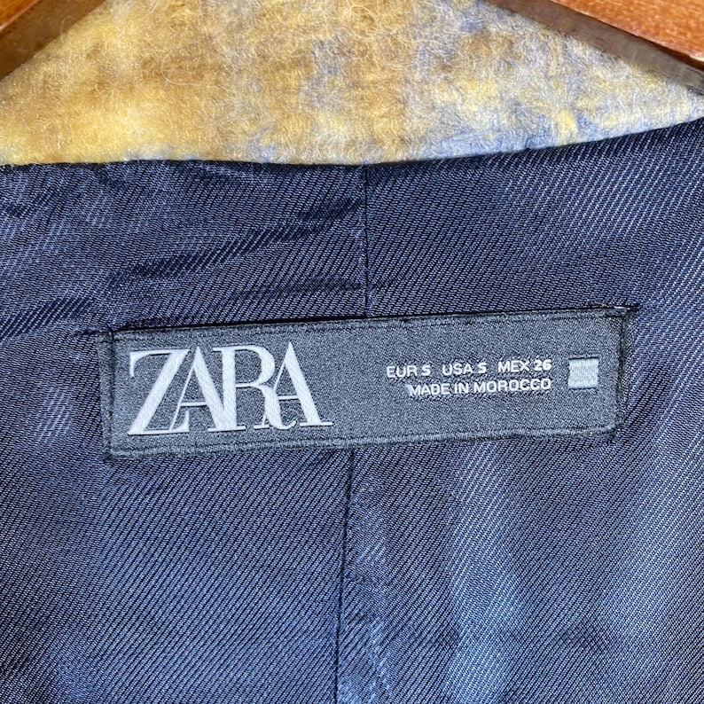 May include: Close-up of a navy blue blazer lining with a ZARA label. The label includes size information and the text "MADE IN MOROCCO". The blazer's interior fabric has a subtle textured pattern.