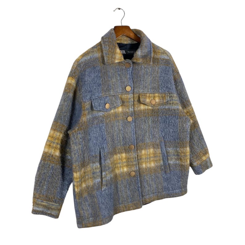 May include: A blue and yellow plaid jacket with a button-up front and two chest pockets. The jacket is made of a soft, fuzzy material and hangs on a wooden hanger.