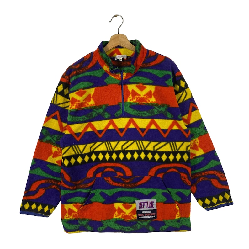 Puede incluir: Un jersey de forro polar colorido con un patr&oacute;n geom&eacute;trico en tonos de rojo, amarillo, verde, azul y negro. El jersey tiene una cremallera y un peque&ntilde;o parche con el texto "NEPTUNE" y "ADVENTURES EXPRESS" en la parte delantera.
