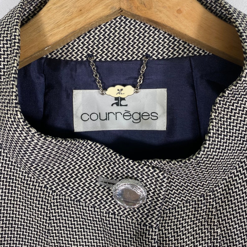 Puede incluir: Una chaqueta con estampado en blanco y negro con un bot&oacute;n plateado y una etiqueta que dice "Courr&egrave;ges" con un peque&ntilde;o logotipo encima del texto.