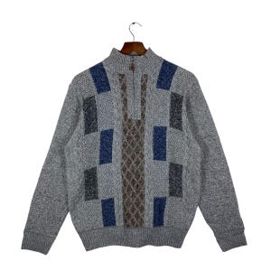 May include: A gray zip-up sweater with a high collar, hanging on a wooden hanger. The sweater features a geometric pattern of blue, gray, and brown squares and diamond shapes. The sweater has long sleeves.