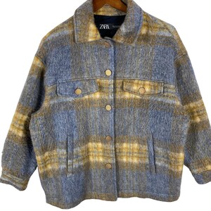 May include: A button-down jacket with a plaid pattern in shades of blue, yellow, and brown. The jacket has two chest pockets and two lower pockets. The brand label "ZARA" is visible on the inside of the collar.