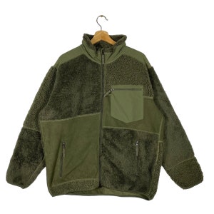 Pode incluir: Casaco de lã polar verde-oliva com gola alta e zíper completo. O casaco apresenta uma mistura de texturas, incluindo painéis lisos e felpudos, e um bolso no peito. Está pendurado num cabide de madeira.