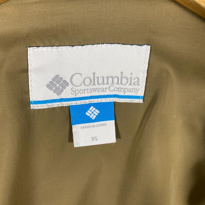 Puede incluir: Una etiqueta blanca con el logotipo y el texto de Columbia Sportswear Company. La etiqueta tambi&eacute;n incluye una etiqueta azul m&aacute;s peque&ntilde;a con el texto "Hecho en China" y "XL".