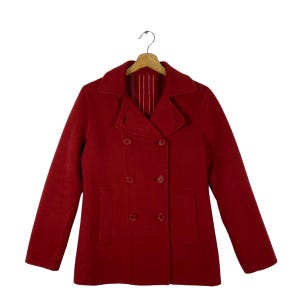 May include: A red double-breasted wool coat with two rows of buttons, a notched collar, and two front pockets. The coat is hanging on a wooden hanger. The coat's color is a deep, rich red.