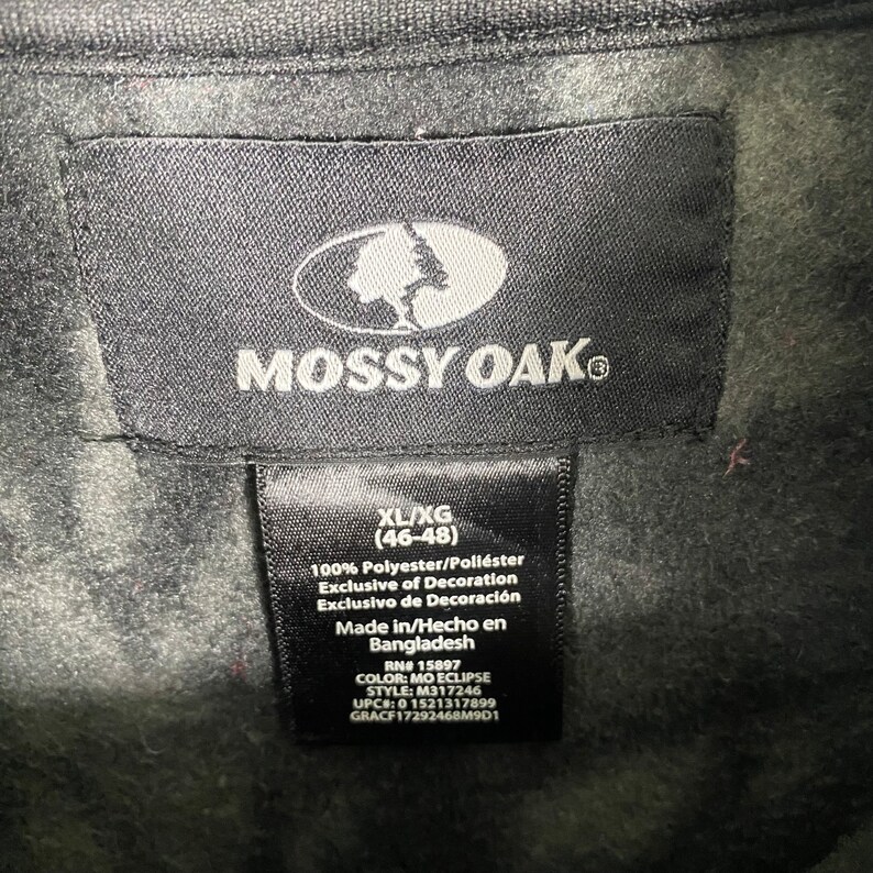 May include: A black Mossy Oak clothing tag with white text. The tag shows the size XL/XG (46-48), fabric content, care instructions, and other details.