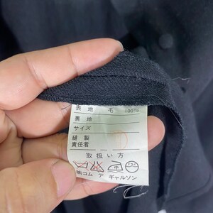 Puede incluir: Una etiqueta blanca con texto negro que detalla el contenido de la tela y las instrucciones de cuidado para una prenda. La etiqueta dice "表地 毛 100% 裏地 サイズ 縫製 責任者 取扱い方 (株)コム デ ギャルソン" que se traduce como "Tejido: 100% Lana, Forro: Talla: Costura: Responsable: Instrucciones de cuidado: (Stock) Comme des Gar&ccedil;ons."