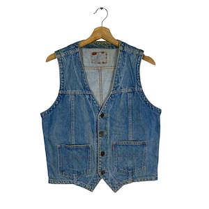 May include: A light blue denim vest with a V-neck and four dark buttons. The vest has two front pockets and is suspended from a wooden hanger. A small red tag is visible on the front pocket.