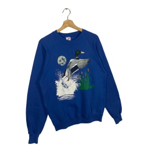 K&ouml;nnte beinhalten: Ein k&ouml;nigsblauer Pullover mit einem grafischen Aufdruck eines Enten, das &uuml;ber einen See mit Schilf fliegt. Die Ente hat einen gr&uuml;nen Kopf und einen wei&szlig;en K&ouml;rper.