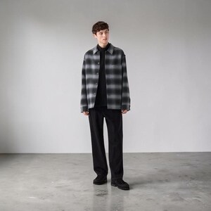 Könnte beinhalten: Ein langärmeliges Hemd mit Knopfleiste und einem grau-schwarzen Ombré-Karomuster, getragen über einem einfarbigen schwarzen Stehkragen-Shirt. Das Outfit wird durch eine einfarbige schwarze Hose und schwarze Schuhe ergänzt, was einen stimmigen Look ergibt.