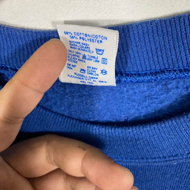 K&ouml;nnte beinhalten: Ein blaues Baumwoll- und Polyester-Sweatshirt-Etikett mit Pflegehinweisen und dem Text "50% Baumwolle/Coton 50% Polyester", "Maschinenw&auml;sche warm - Farben separat waschen", "Nur nicht-chlorhaltiges Bleichmittel verwenden, wenn n&ouml;tig", "Trocknen bei niedriger Temperatur", "Nicht b&uuml;geln", "Chemische Reinigung oder Reinigung des Emblems", "Russell Corp. Alexander City, AL. 35019", "WPL 7232".