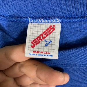 K&ouml;nnte beinhalten: Ein blaues Jerzees-Markensweatshirt-Etikett mit wei&szlig;em Hintergrund. Das Etikett hat ein Gittermuster und den Text "Jerzees" in Rot. Die Gr&ouml;&szlig;e ist mit "L 42-44" gekennzeichnet und das Etikett lautet "Made in U.S.A. For care & content see reverse."