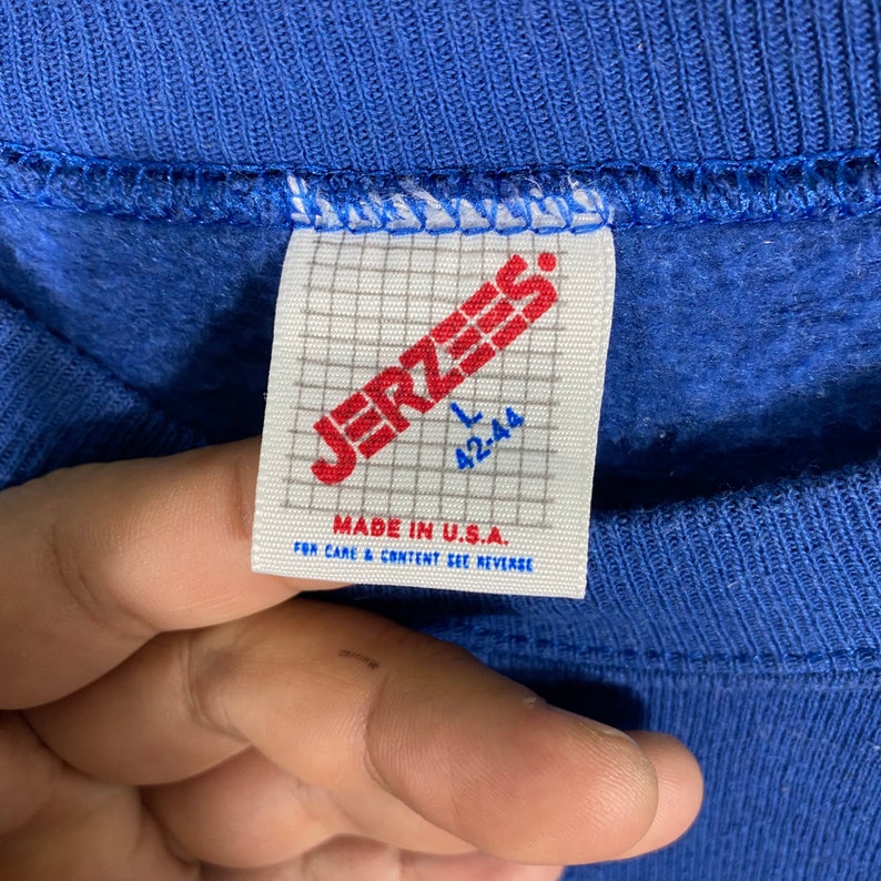 K&ouml;nnte beinhalten: Ein blaues Jerzees-Markensweatshirt-Etikett mit wei&szlig;em Hintergrund. Das Etikett hat ein Gittermuster und den Text "Jerzees" in Rot. Die Gr&ouml;&szlig;e ist mit "L 42-44" gekennzeichnet und das Etikett lautet "Made in U.S.A. For care & content see reverse."