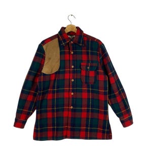 May include: A long-sleeved plaid shirt in red, green, and yellow, with a brown suede shoulder patch and a chest pocket. The shirt is button-down and hangs from a wooden hanger.