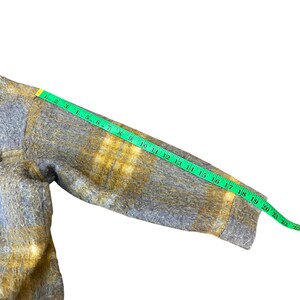 May include: A gray and yellow plaid jacket sleeve is measured with a green measuring tape. The sleeve is approximately 22 inches long. The jacket has a fuzzy texture and a buttoned cuff.
