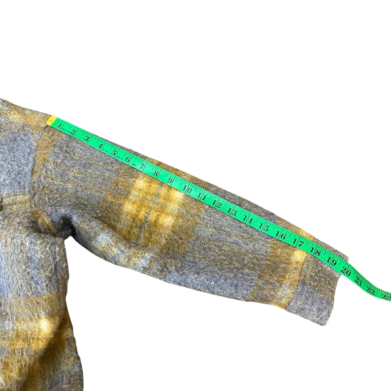 May include: A gray and yellow plaid jacket sleeve is measured with a green measuring tape. The sleeve is approximately 22 inches long. The jacket has a fuzzy texture and a buttoned cuff.